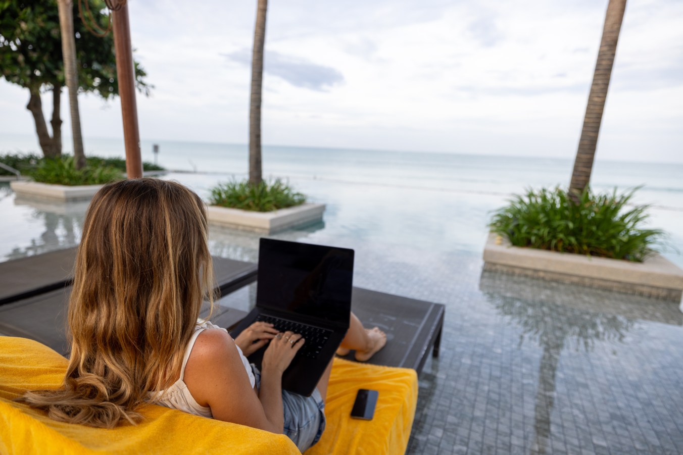 A woman works on her laptop while lounging by an infinity pool, overlooking the ocean on a serene tropical day.