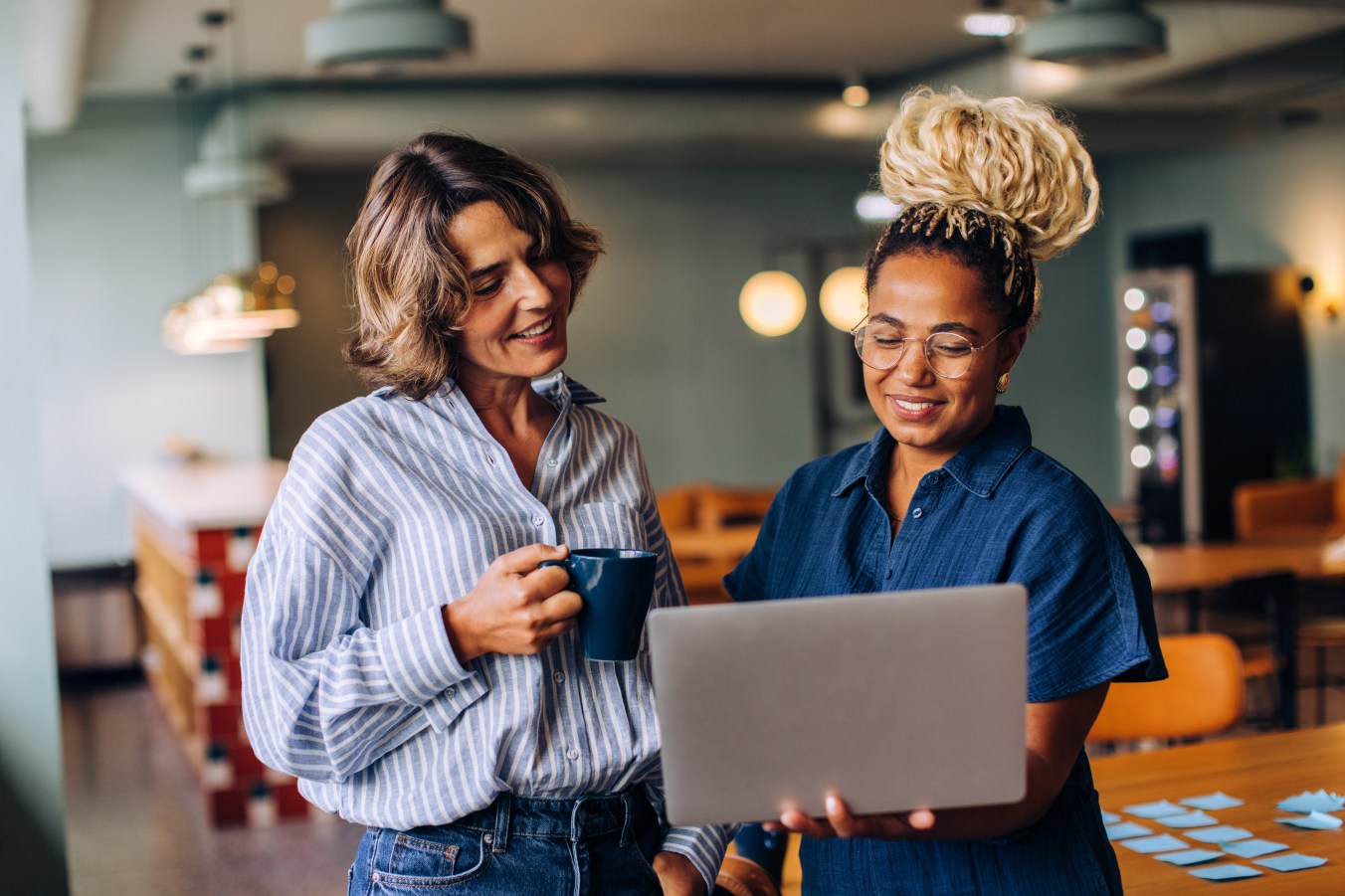Two women are standing in a bright office, holding a laptop and coffee while smiling and engaged in a discussion, showcasing a cheerful interaction during a professional or casual working environment.